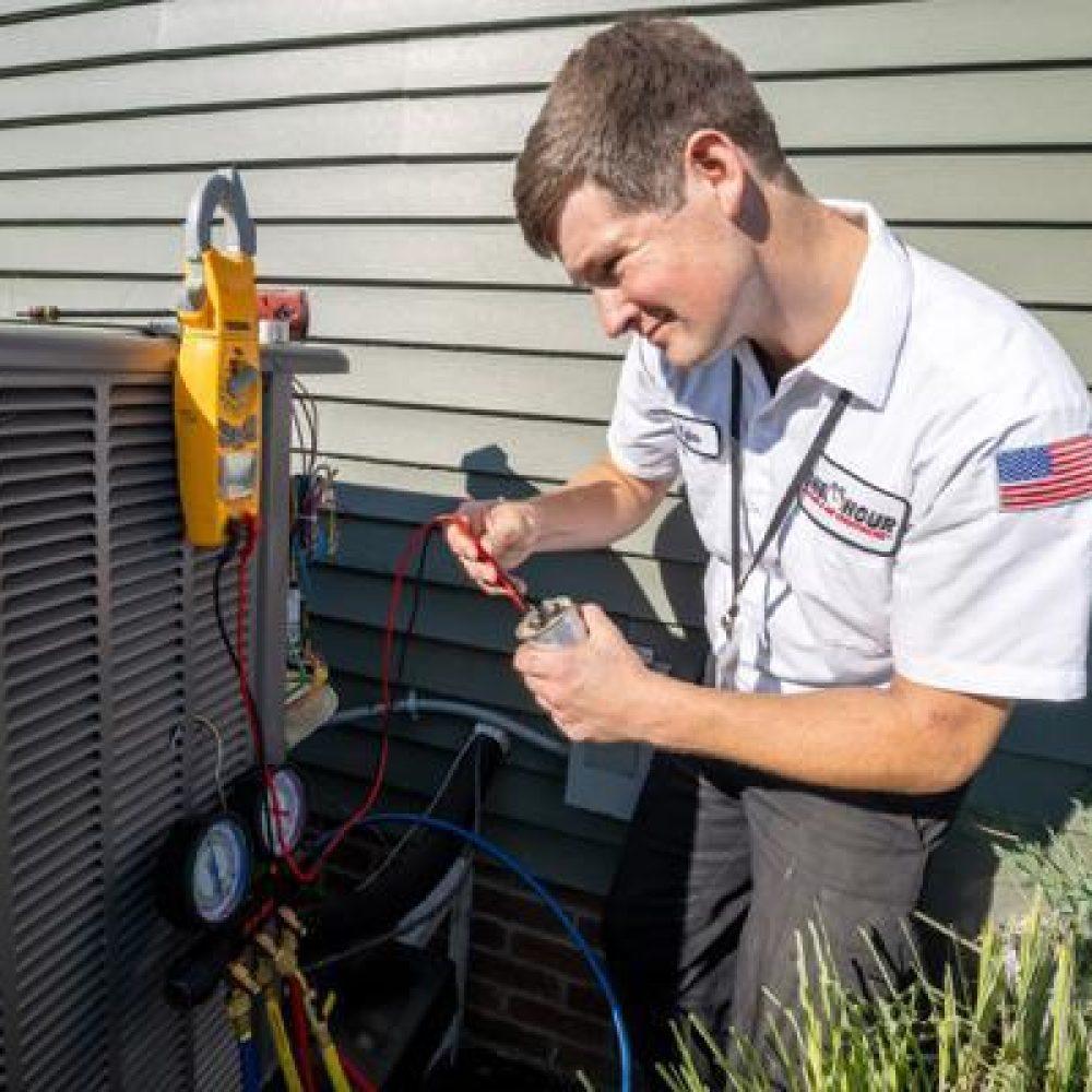 A technician in a white uniform with an American flag patch repairs an outdoor air conditioning unit, using tools and gauges, near a house with green siding.