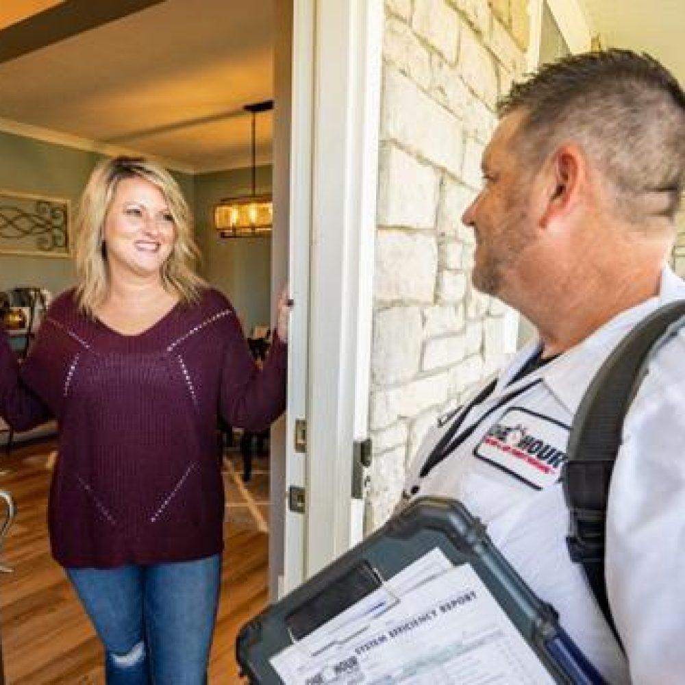 A woman stands at her front door smiling at a technician, who is holding a clipboard and wearing a uniform with an American flag patch and a 24 Hour logo. They appear to be having a friendly conversation.