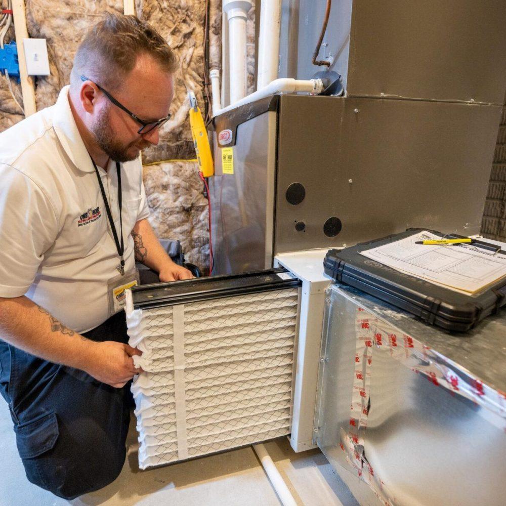 A technician wearing glasses and a white shirt is kneeling and replacing an air filter in a furnace unit inside a basement. A black folder with documents rests on top of the ductwork.